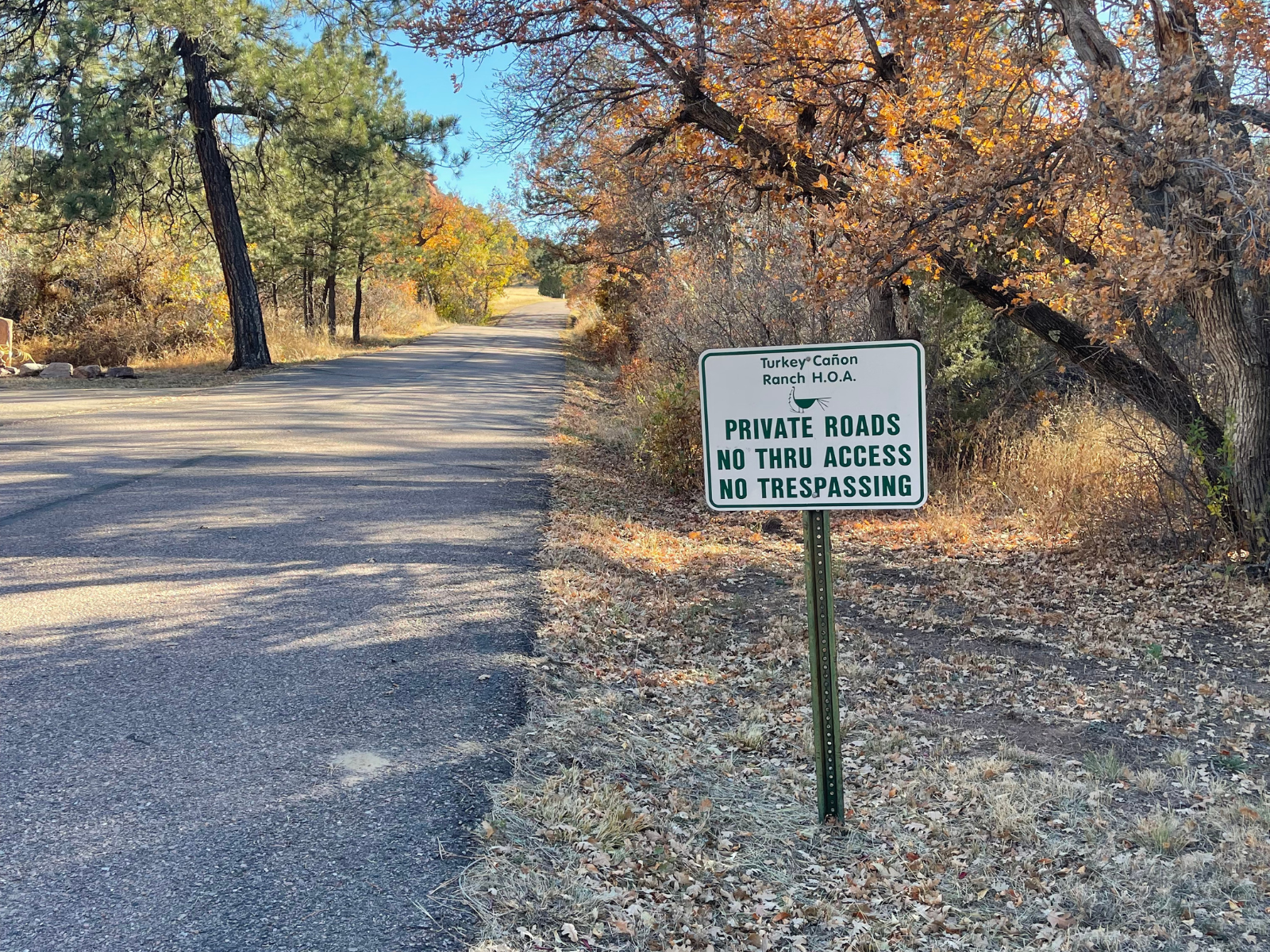A white roadside sign reading “Private Roads No Thru Access” beside a paved road lined with autumn aspen trees.